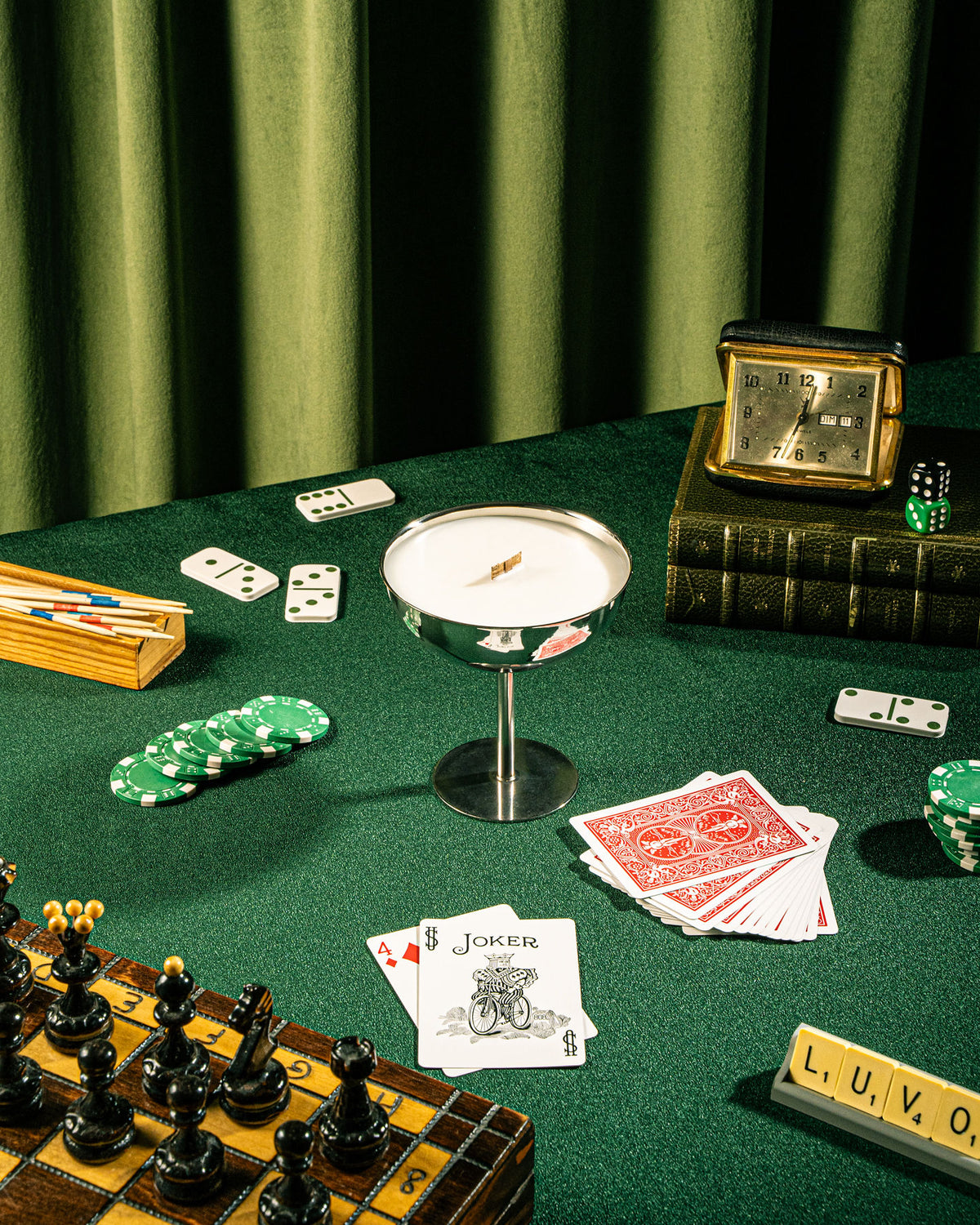 Lit Célébration champagne cup candle on a green game table with playing cards, dominoes, chess set and LUVO letter tiles