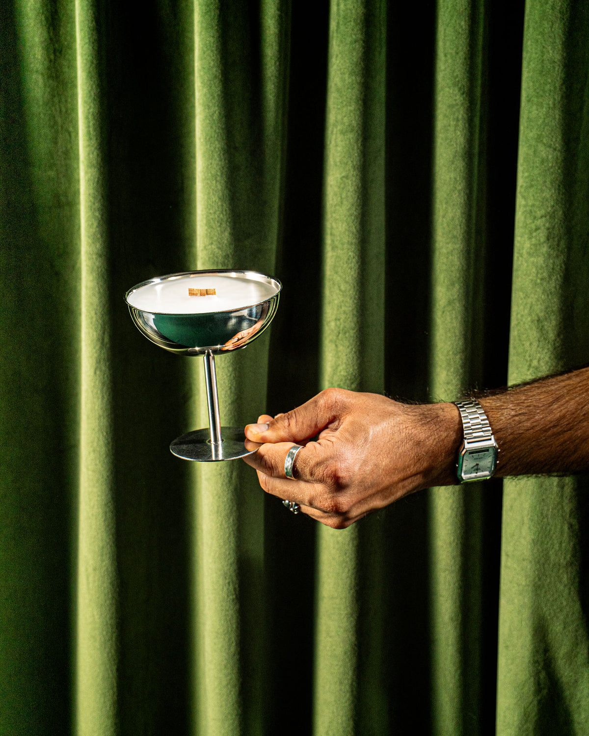 Hand holding the LUVO Célébration wooden wick candle in a reusable stainless steel champagne cup against green velvet